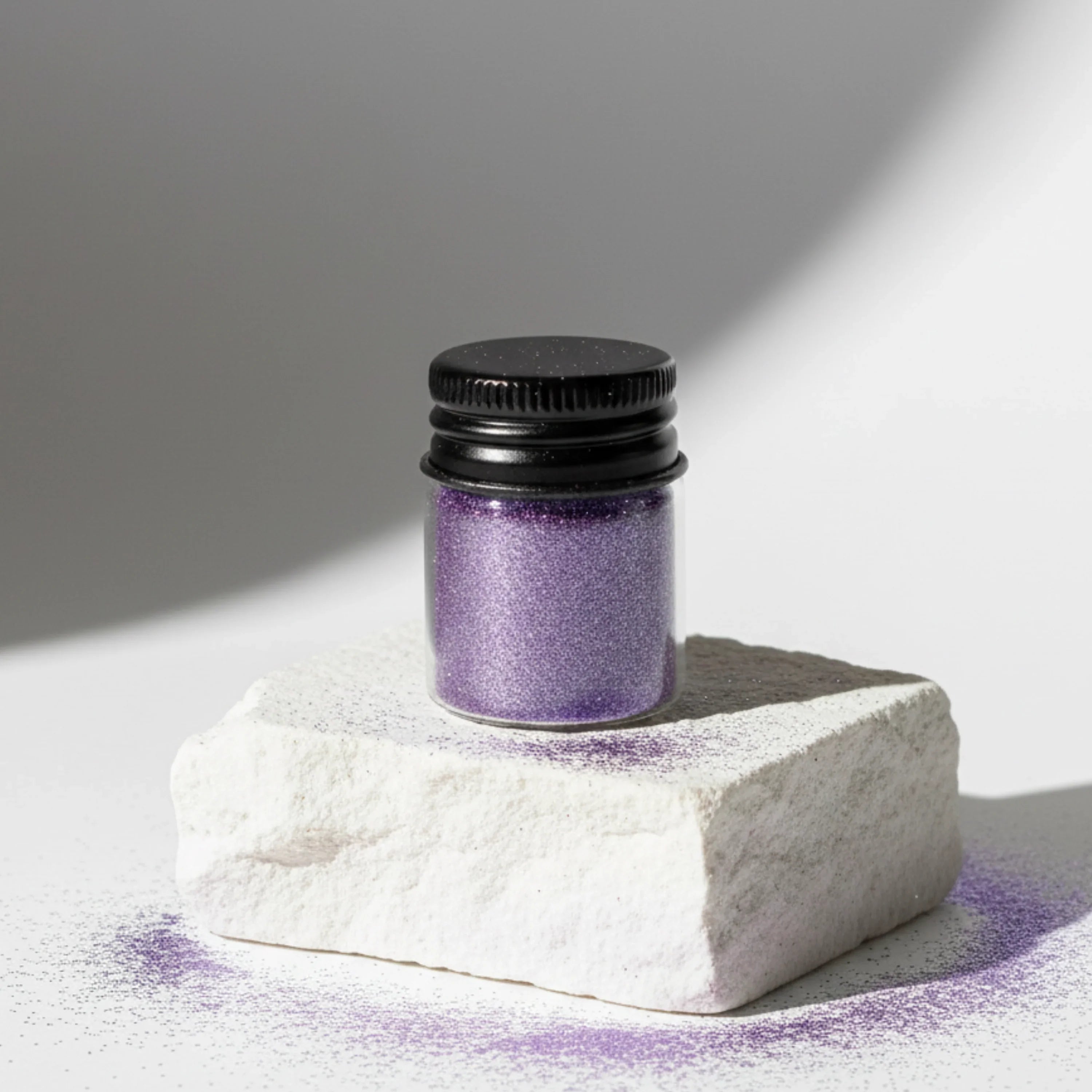 Small jar of purple glitter with a black lid placed on a white textured stone, with scattered glitter around it.